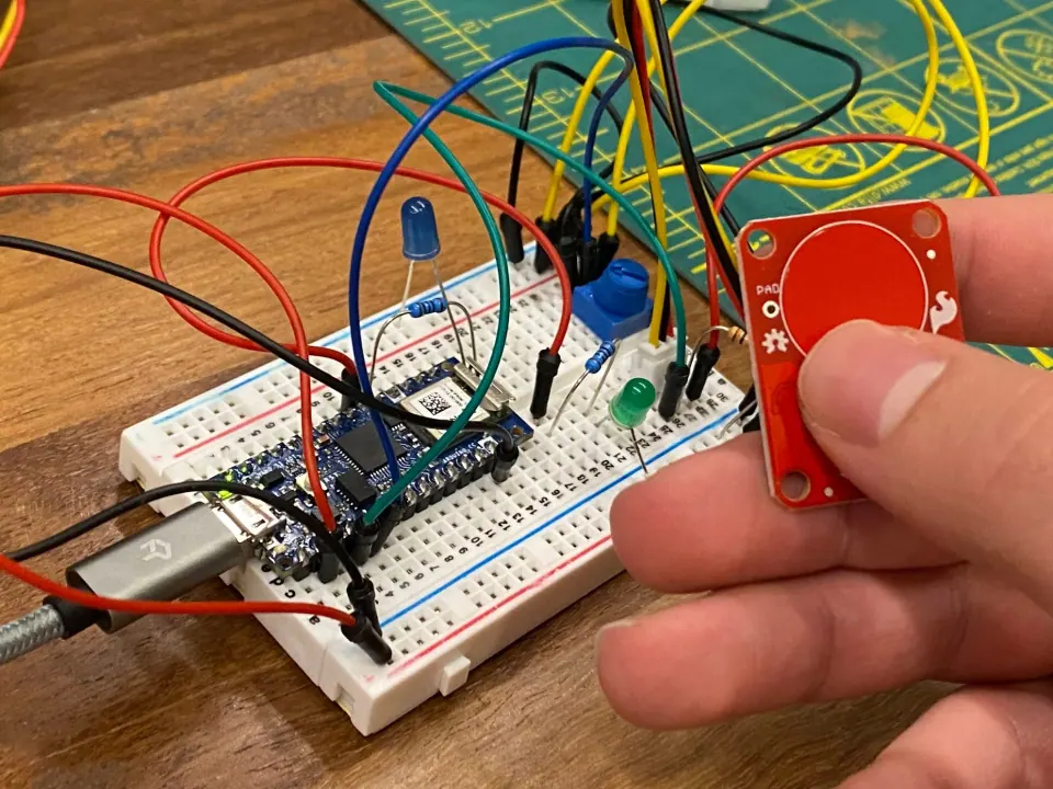 A hand holding a red capacitive touch sensor board next to the breadboard circuit with its blue and green LEDs