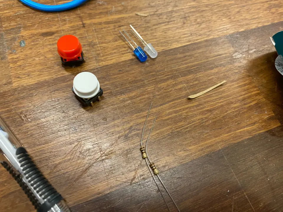 A red button, a white button, blue and white LEDs and a resistor laid out on a wooden bench
