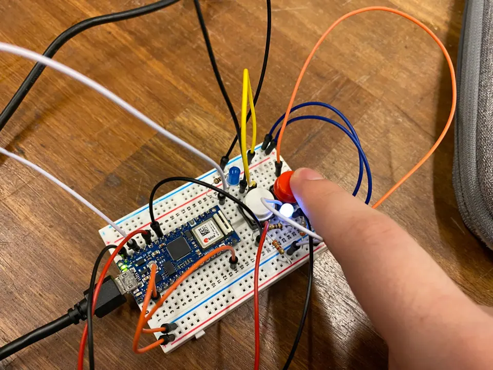 A finger pressing the red pushbutton while a white LED lights on the breadboard