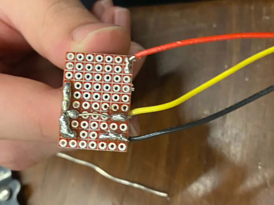 Underside of the button protoboard held between fingers, showing small solder traces and red, yellow and black wires