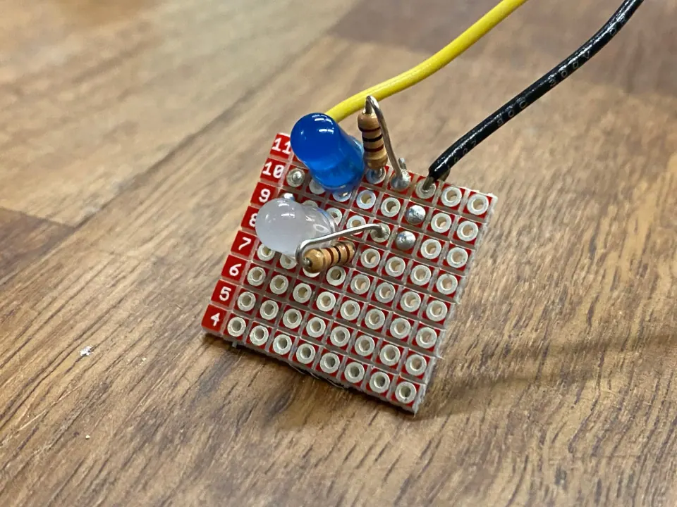 Top of the LED protoboard with a blue LED, a white LED and a resistor soldered in, and yellow and black wires