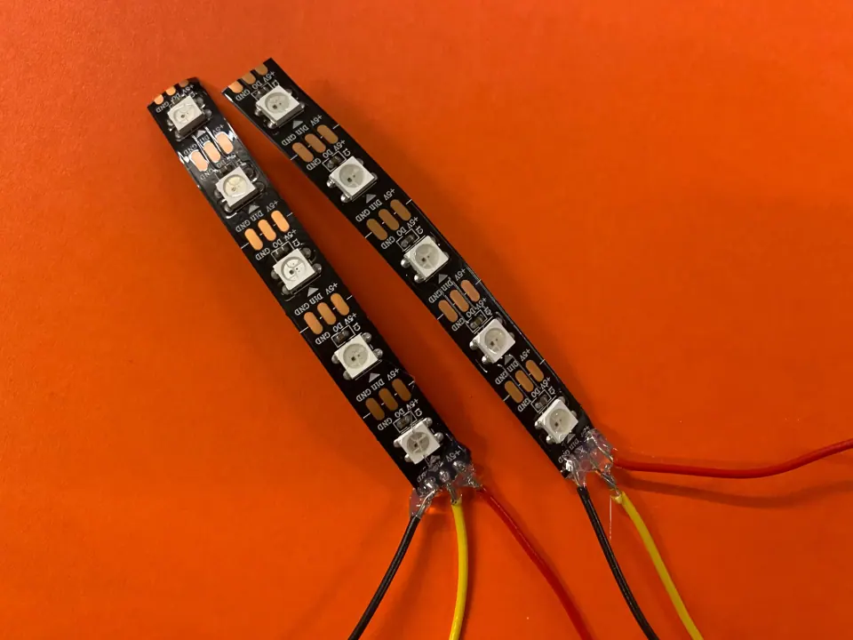 Two black WS2812 NeoPixel LED strips with soldered red, yellow and black wires on an orange background