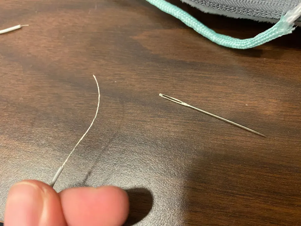 A strand of thread held between fingers next to a large sewing needle on a dark wooden table