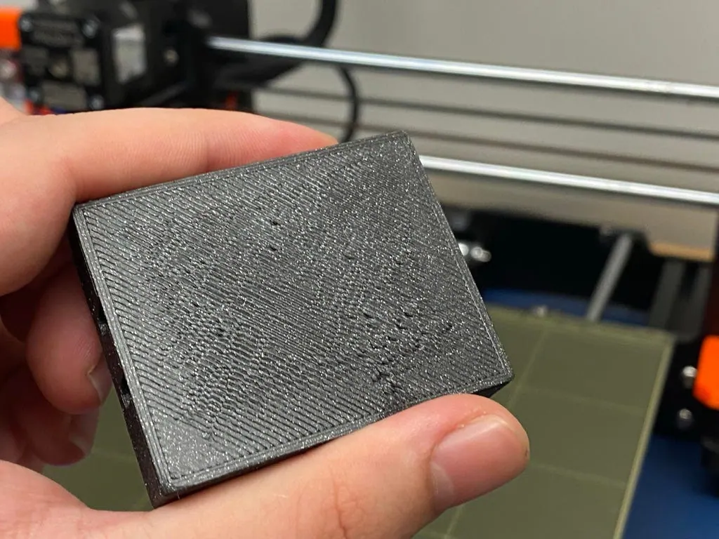 Hand holding another printed housing part with visible layer lines, in front of a Prusa i3 MK3 printer bed
