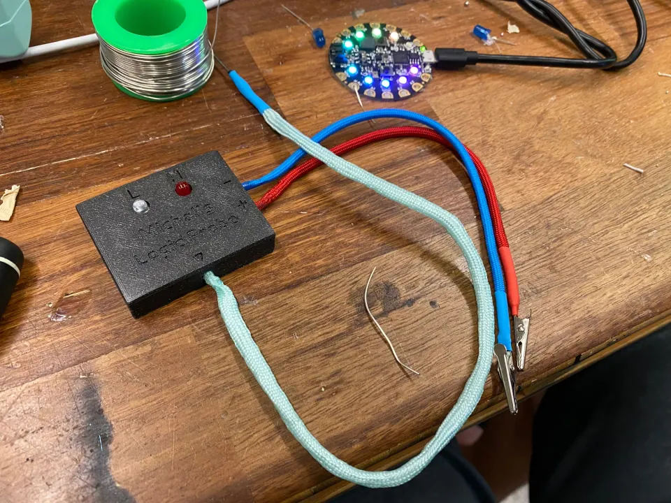 Logic probe on the workbench with alligator clips on its blue and red cords, beside a Circuit Playground board and a spool of solder