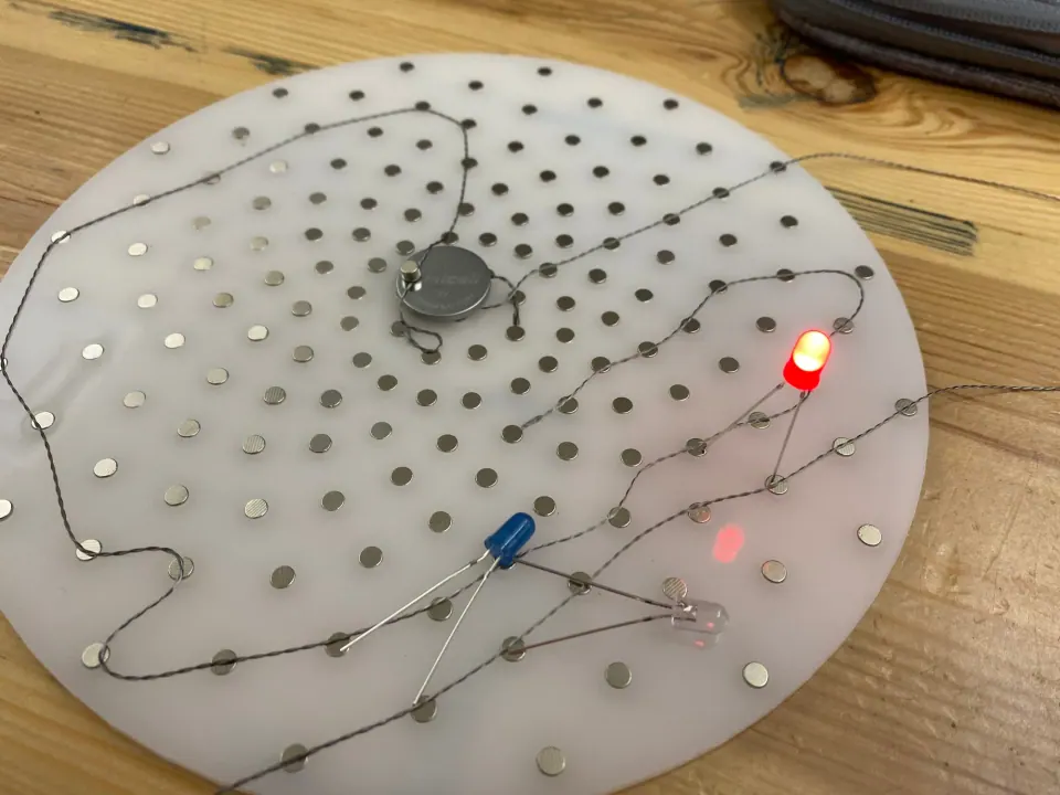 ThreadBoard, a white acrylic disc studded with magnets and stitched with magnetic thread, with a coin cell battery and a red LED lit and blue and white LEDs unlit
