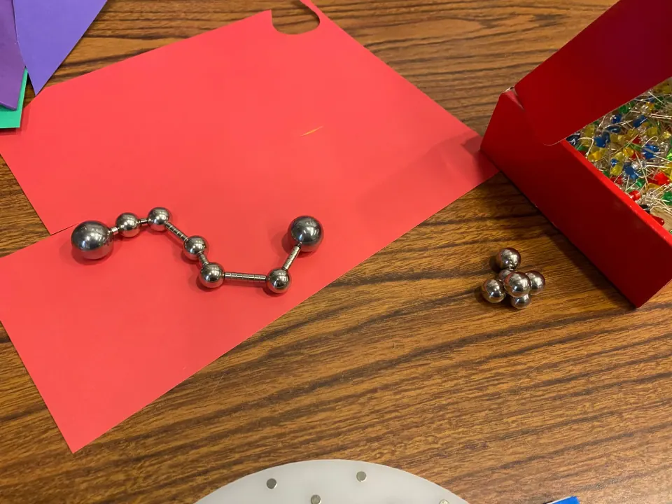Chain of small steel balls and magnets bent into a constellation shape on red paper, beside a pile of balls and a box of LEDs