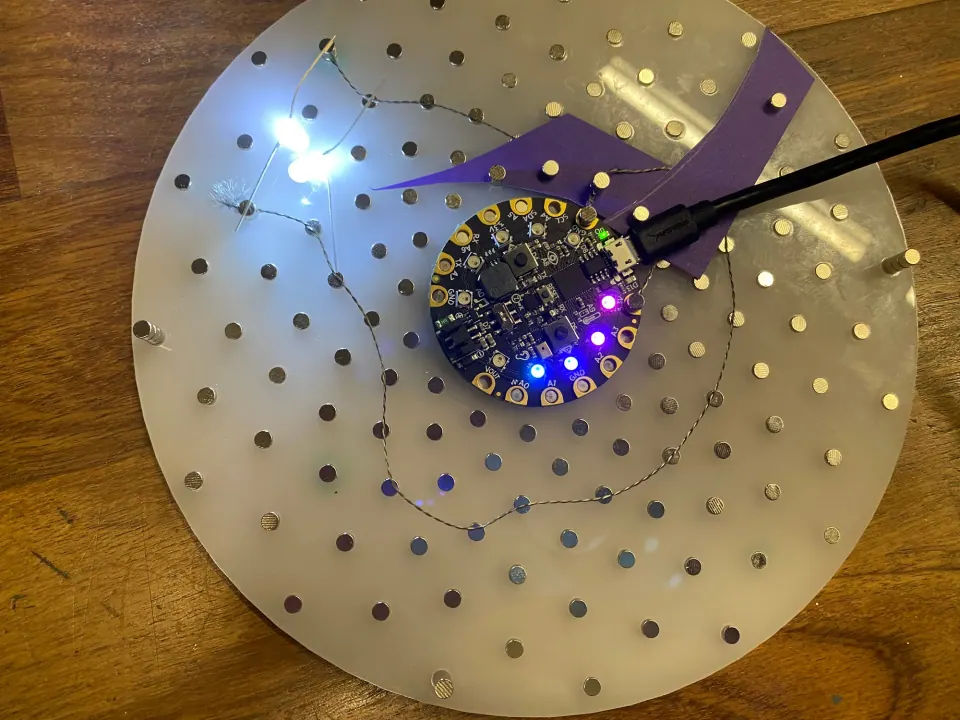ThreadBoard with an Adafruit Circuit Playground and purple paper in the centre and a white LED lit, the prototype circuit for the first contact pads