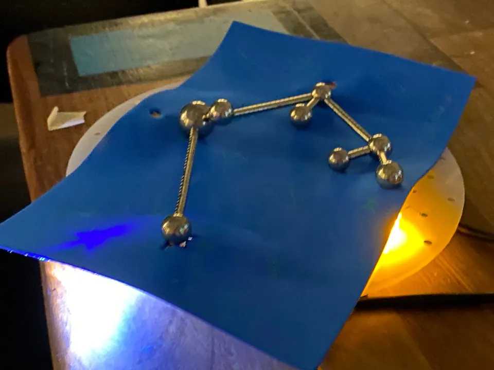 Blue foam sheet over the lit board with steel balls and bars joined into a constellation shape, with yellow and white light bleeding through