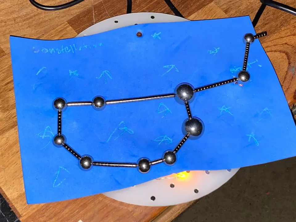 Constellation of steel balls and magnetic bars shaped like the Big Dipper on blue foam with hand-drawn stars and handwriting, with a yellow glow underneath