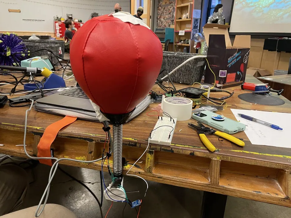 Red punching bag on a spring mount with an Arduino and wires hanging below it, on a workshop table with tools