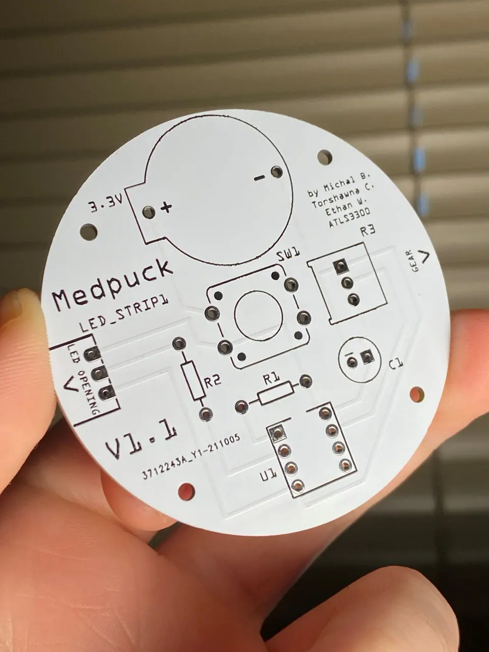 Hand holding the bare round MedPuck PCB, with white silkscreen labels and a coin cell outline