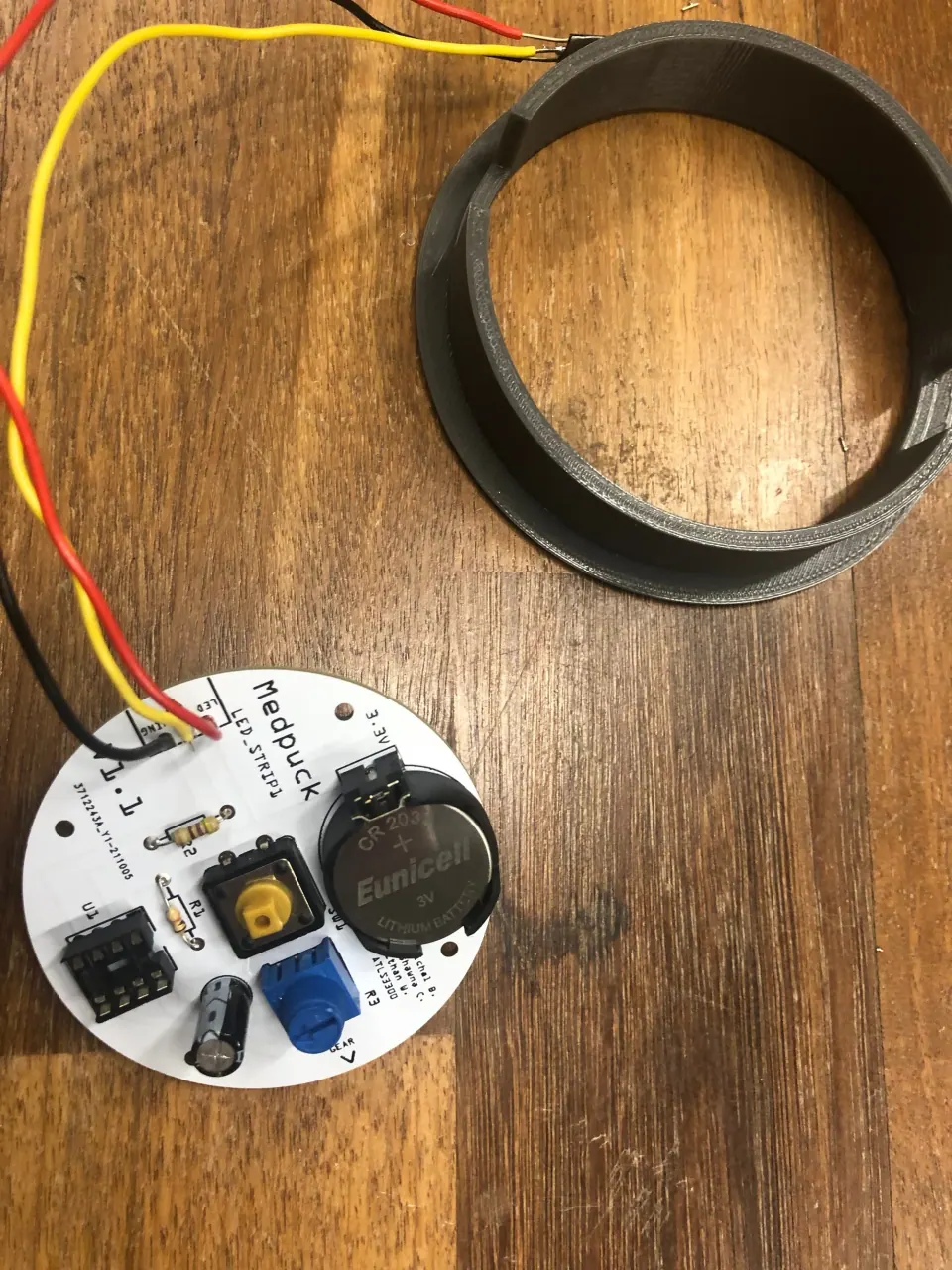 Soldered MedPuck PCB with a coin cell, trim pot and pushbutton and yellow and red wires, beside the grey printed ring