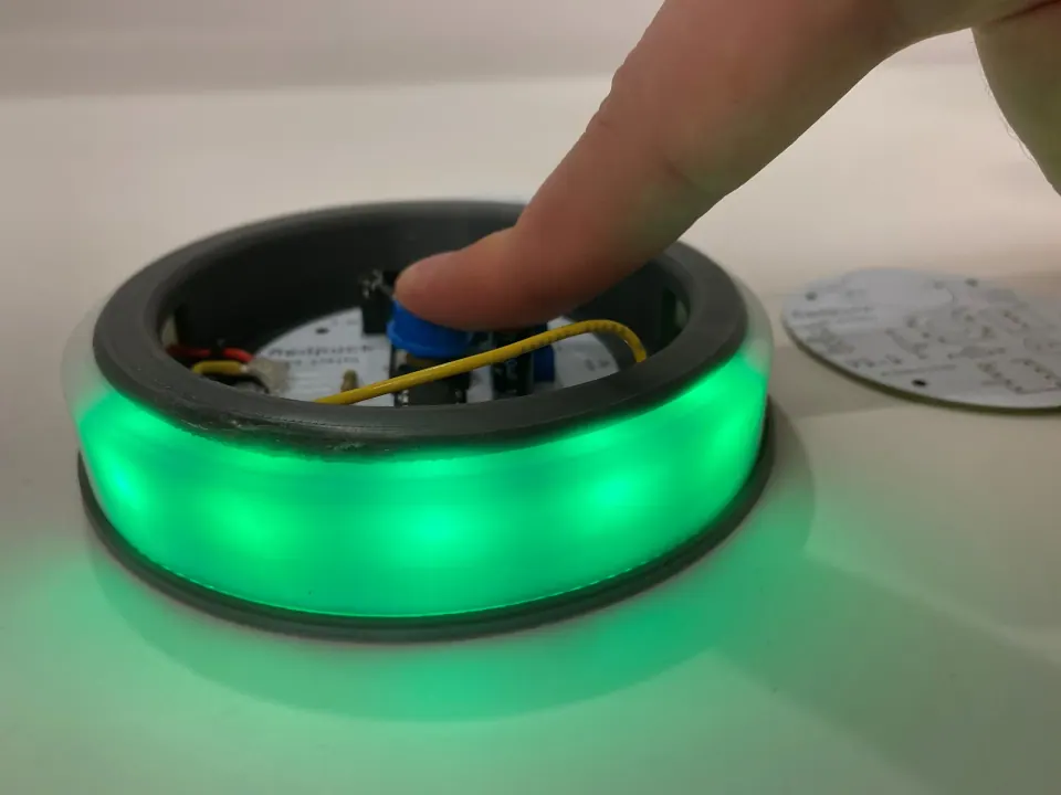 Fingertip pressing the blue pushbutton in the assembled MedPuck while the LED ring glows green