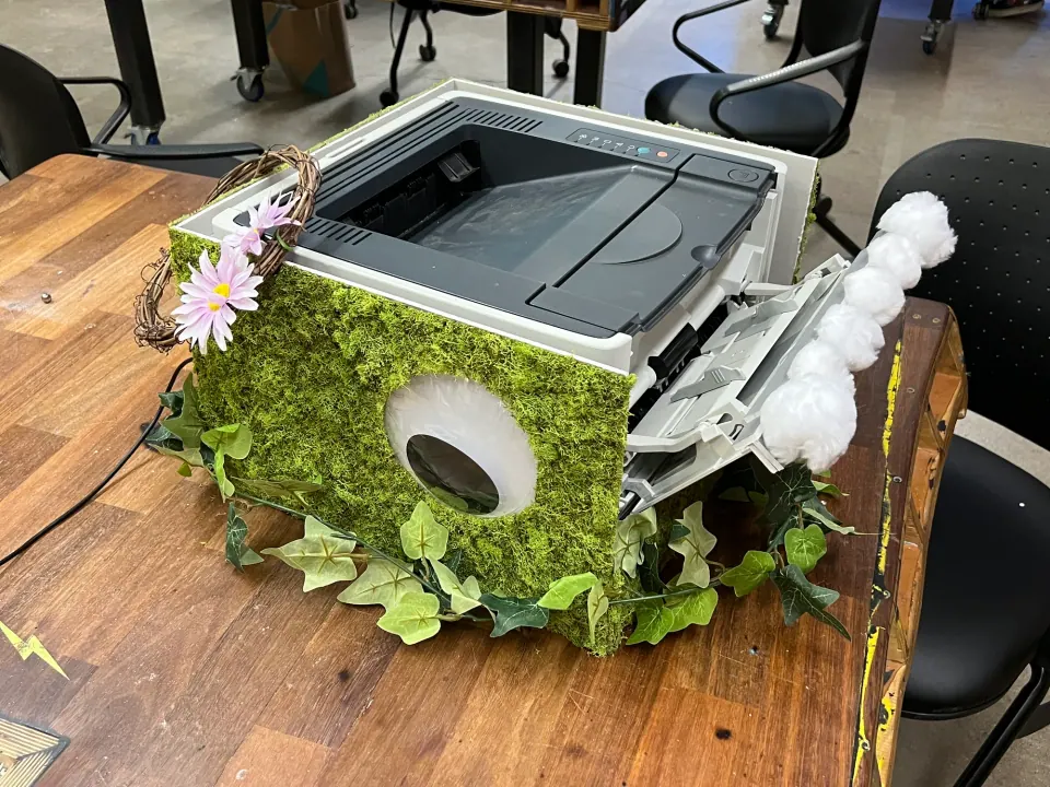 A printer wrapped in moss and flowers, opened up as the game enclosure